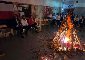 Ognisko, a w tle widoczni siedzący: Pani dyrektor, wicedyrekcja i gość oraz młodzież, dekoracje patriotyczne