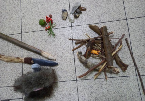 Patyki złożone w ognisko, obok narzędzia kamienne, jagody, owoce i orzechy, oszczep i włócznia, zwierzę futerkowe