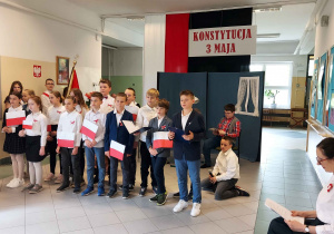 Na scenie, na tle polskiej flagi i napisu Konstytucja 3 maja widać chór (klasa 4b i innych uczniów z 4a, 5b, 6a z biało-czerwonymi chorągiewkami), z tyłu poczet sztandarowy i aktorzy z 4a i 4b. Za nimi wychowawczyni 4a, nauczyciel informatyki.